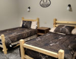 Photo of two beds made up in hunting-themed sheets separated by a night table with the MWA logo above.